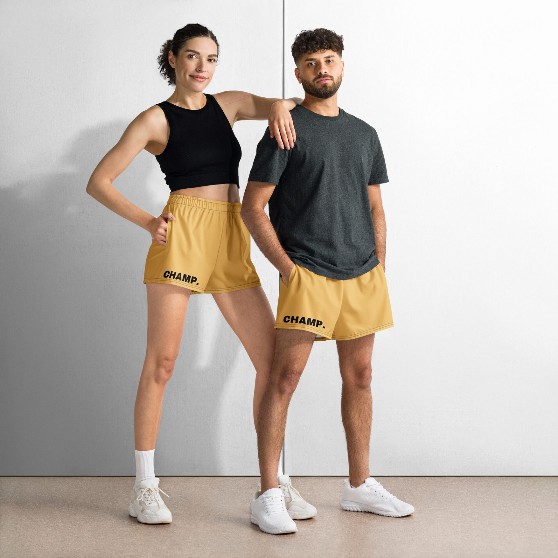 A woman in a black tank top and CHAMP Golden Athletic Shorts stands confidently alongside a man in a gray t-shirt and matching shorts. CHAMP. is emblazoned on their golden shorts, as both have hands in pockets, sporting white sneakers against a two-tone wall. A woman in a black tank top and CHAMP Golden Athletic Shorts stands confidently alongside a man in a gray t-shirt and matching shorts. CHAMP. is emblazoned on their golden shorts, as both have hands in pockets, sporting white sneakers against a two-tone wall.