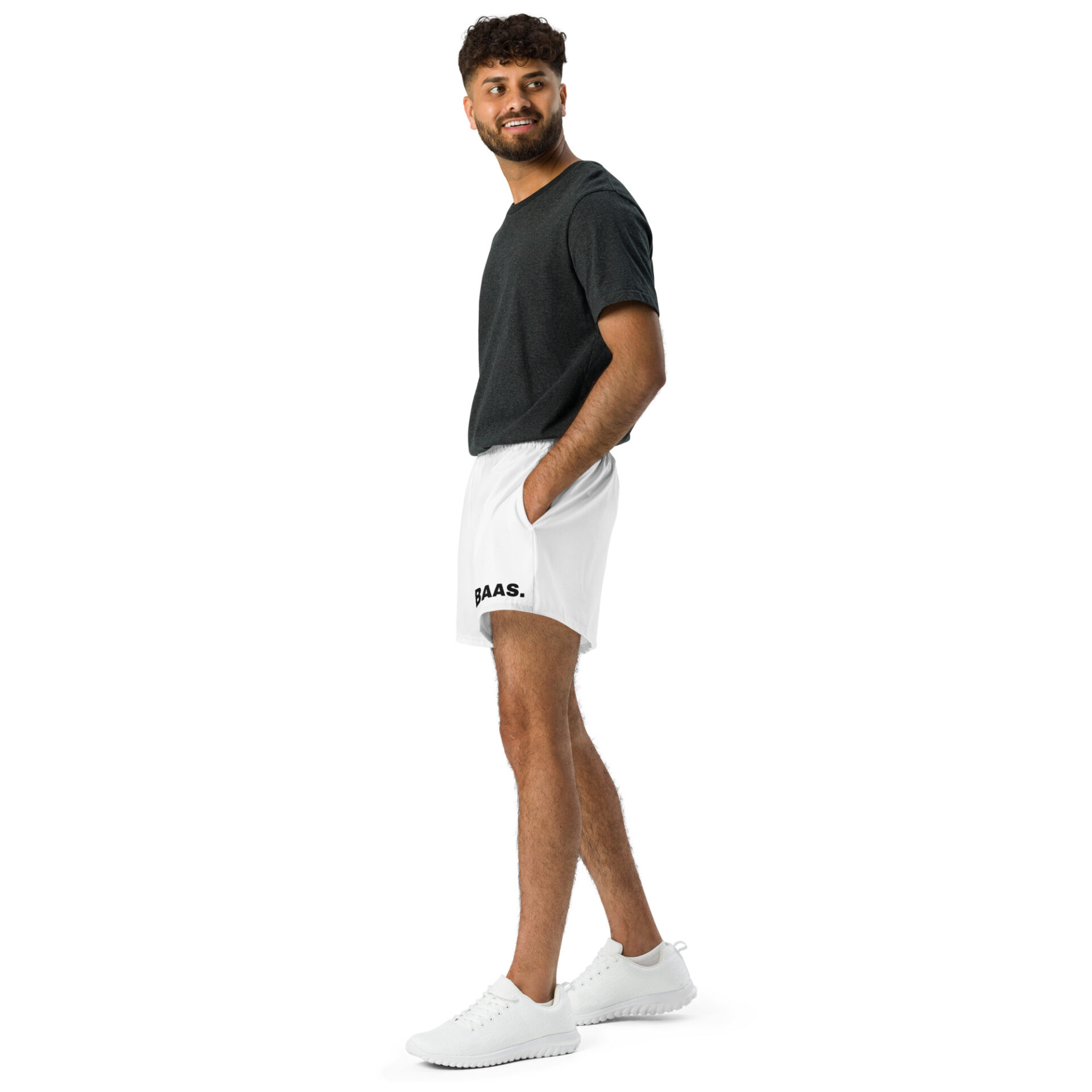 A bearded man smiles in dark gray T-shirt and All-Over Print Unisex Athletic Shorts with BAAS, wearing white sneakers. His hands are in his pockets, and he stands slightly angled against a white background.