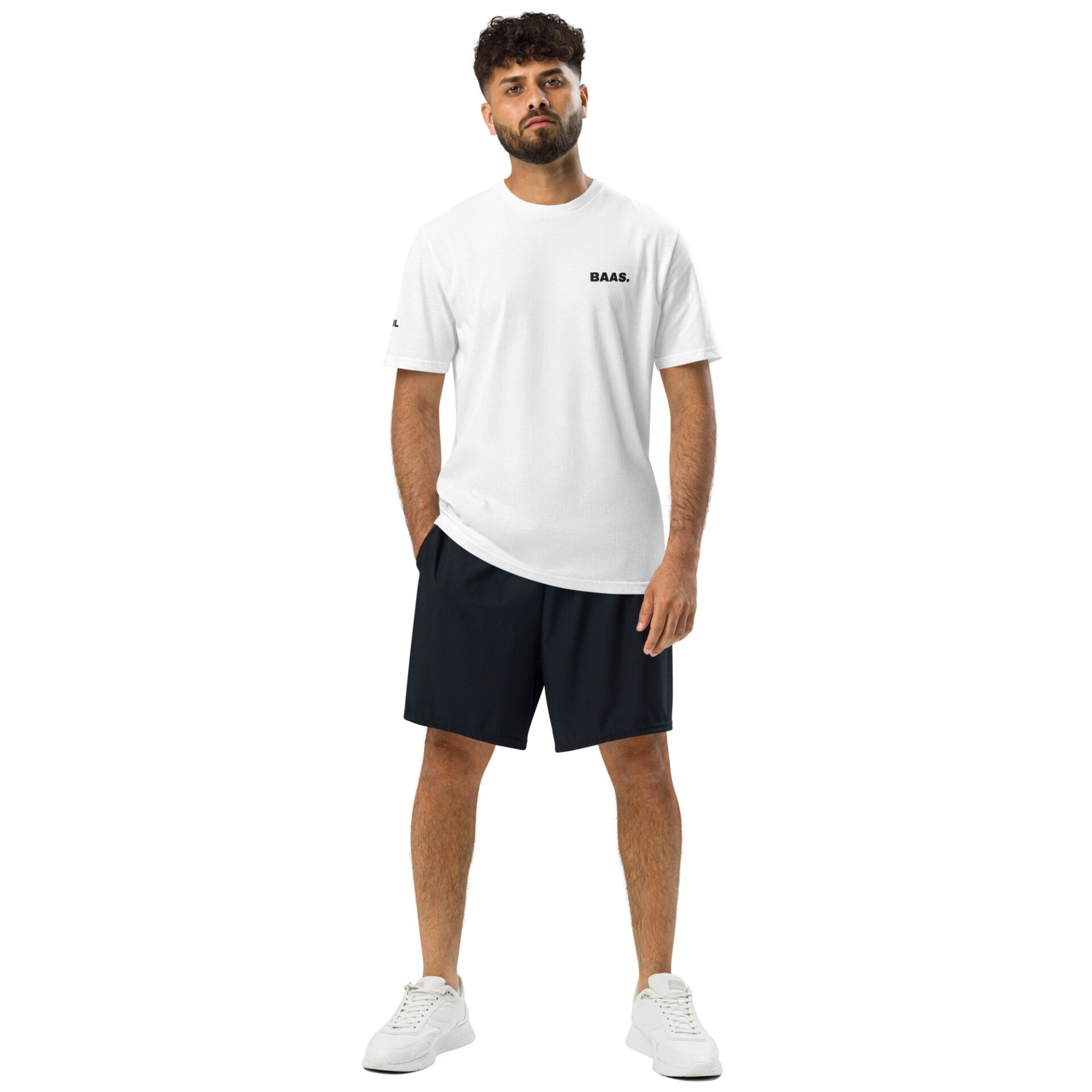 A man with curly hair and a beard stands against a white background, wearing an Under ArmourⓇ athletic t-shirt with BAAS. on the chest, black shorts, and white sneakers, hands by his sides. A man with curly hair and a beard stands against a white background, wearing an Under ArmourⓇ athletic t-shirt with BAAS. on the chest, black shorts, and white sneakers, hands by his sides.